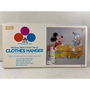 May include: A vintage Dolly Toy Walt Disney "Mickey & Donald" Tidee-Up Clothes Hanger. The hanger features Mickey Mouse and Donald Duck characters, a wooden beam design, and colorful accents. The box includes text about the product.