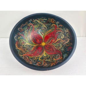 May include: A round, hand-painted wooden bowl with a dark blue exterior. The interior features a vibrant floral design in red, yellow, and green against a black background. The bowl is a decorative piece.