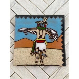 Könnte beinhalten: Quadratische Keramikfliese mit einem indianischen Tänzer in einem farbenfrohen Design. Der Tänzer hat ausgestreckte Arme mit weißen Federflügeln, eine grüne Maske und einen weißen Rock. Der Hintergrund umfasst einen blauen Himmel, eine beige Wüste und einen schwarzen Rand.