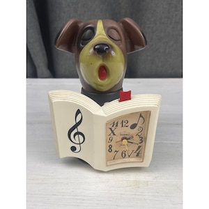 May include: A brown and tan dog figurine with a closed-eye expression and open mouth, positioned behind an off-white open book. The book features a black treble clef on the left page and a clock face with the brand name SUNKO on the right page.