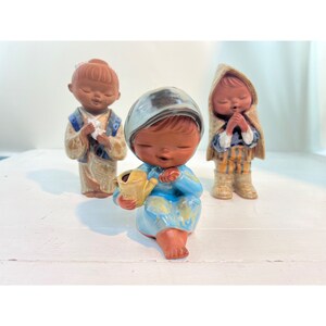 Może przedstawiać: Trzy ceramiczne figurki z zamkniętymi oczami. Jedna figurka siedzi i trzyma żółtą konewkę, ma na sobie niebieską sukienkę i niebiesko-srebrny kapelusz. Pozostałe dwie figurki stoją, jedna w kimonie, a druga w kapturze.