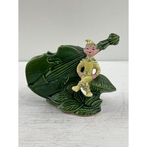 May include: A green ceramic figurine of a violin with a seated elf. The violin is dark green with carved details. The elf is wearing a yellow outfit with a pointed hat and is sitting on a mushroom.