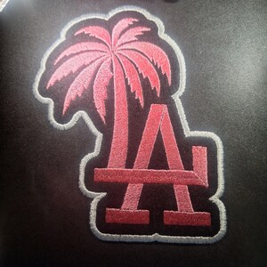 May include: Embroidered patch featuring a pink palm tree and the letter "A" in a stylized design. The patch has a black background with a white border. The patch is likely for apparel or accessories.