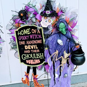 May include: A Halloween wreath featuring a purple witch, a black cauldron, a skeleton bat, and a sign that reads "Home of a spooky witch, one handsome devil and their ghoulish goblins".