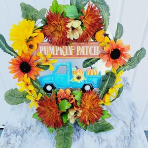 Può includere: Una ghirlanda autunnale con un camioncino blu che trasporta zucche e un cartello con la scritta "Pumpkin Patch". La ghirlanda è decorata con girasoli, fiori arancioni e gialli e foglie verdi.