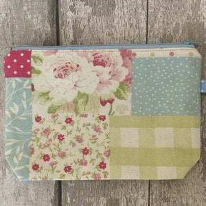 May include: A patchwork fabric zippered pouch with a floral design in shades of pink, green, and blue. The pouch has a light blue zipper and a light blue fabric pull tab.