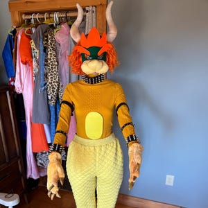 May include: A full-body costume of a Bowser character, featuring a yellow quilted jumpsuit and matching long-sleeved top. The costume includes a Bowser mask with horns, a spiked collar, and clawed gloves and slippers. The costume is displayed on a mannequin.