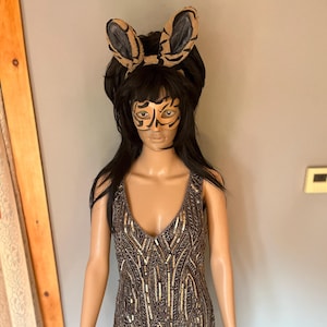 May include: A brown and gold sequined dress with a V-neck, displayed on a mannequin. The dress has a geometric pattern. The mannequin is wearing a black wig, tiger ears, and face paint. The dress is a mini dress.