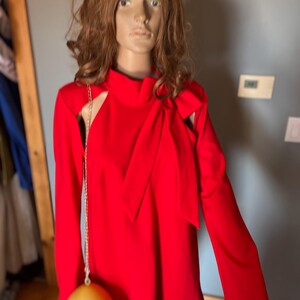 May include: A vibrant red dress with long sleeves and a high neckline, featuring cut-out shoulder details and a bow accent. A gold chain bag with a yellow egg-shaped body and the word "BAD" is hanging from the dress.