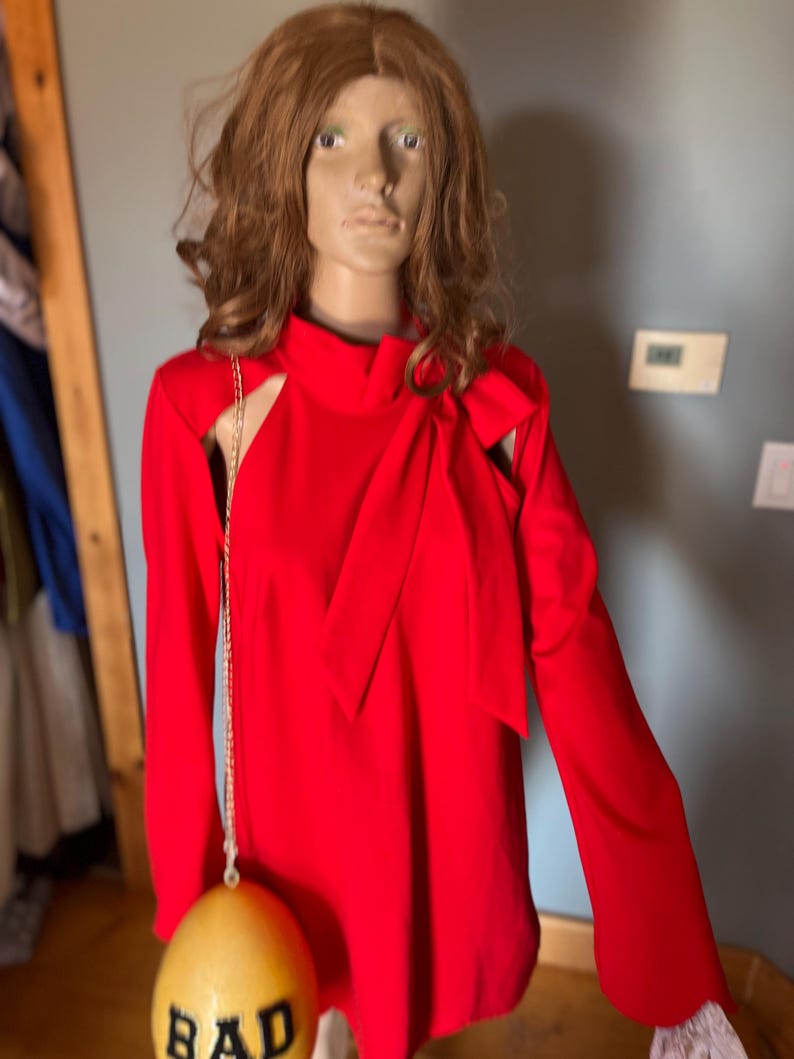May include: A vibrant red dress with long sleeves and a high neckline, featuring cut-out shoulder details and a bow accent. A gold chain bag with a yellow egg-shaped body and the word "BAD" is hanging from the dress.