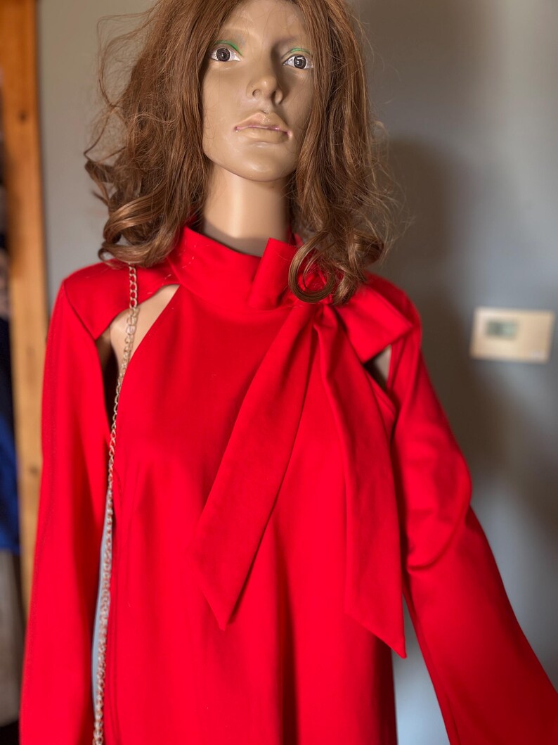 May include: A striking red dress with long sleeves and shoulder cutouts. The dress has a high neckline and a large bow detail. A gold chain strap is visible. The dress is displayed on a mannequin.