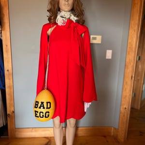 May include: A red dress with long sleeves and a white lace collar is displayed on a mannequin. A yellow egg-shaped purse with the words "BAD EGG" in black is hanging from the mannequin's shoulder.