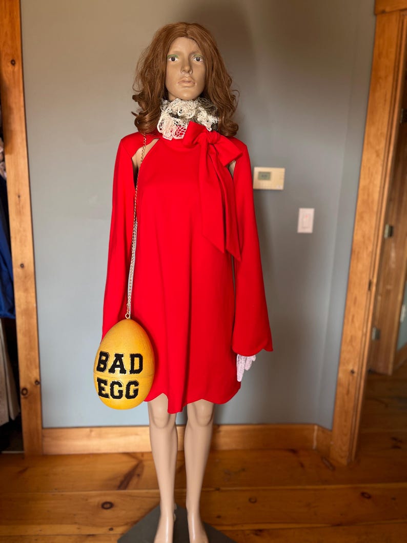 May include: A red dress with long sleeves and a white lace collar is displayed on a mannequin. A yellow egg-shaped purse with the words "BAD EGG" in black is hanging from the mannequin's shoulder.