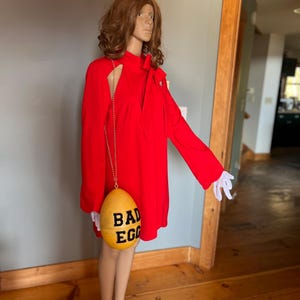 May include: A vibrant red dress with long sleeves and a distinctive shoulder cut-out. A yellow egg-shaped purse, inscribed with "BAD EGG" in black, hangs from a gold chain. The mannequin is wearing white gloves.