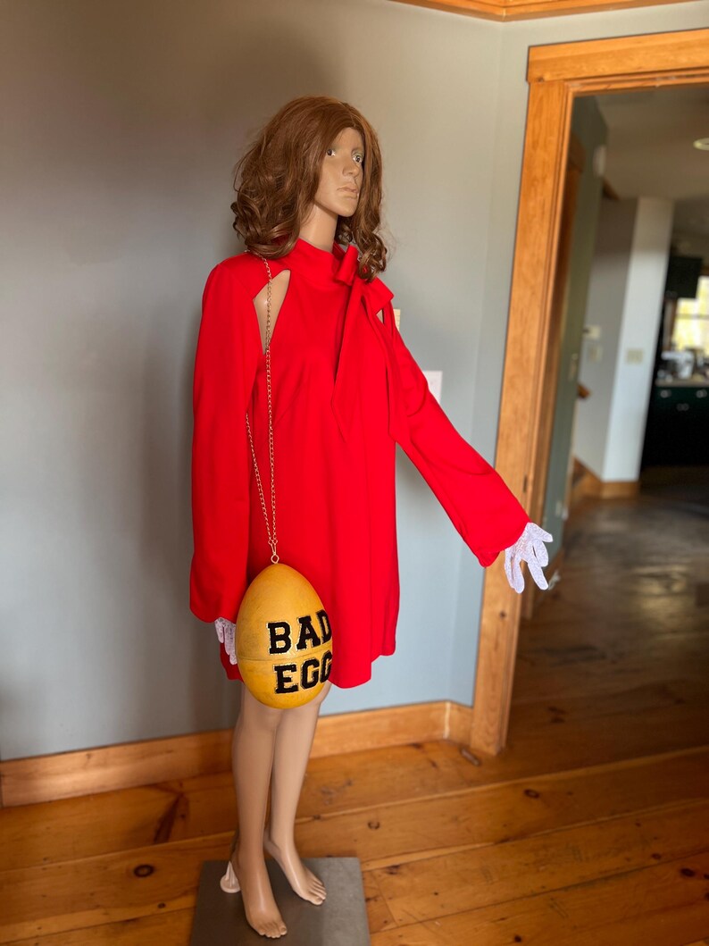May include: A vibrant red dress with long sleeves and a distinctive shoulder cut-out. A yellow egg-shaped purse, inscribed with "BAD EGG" in black, hangs from a gold chain. The mannequin is wearing white gloves.