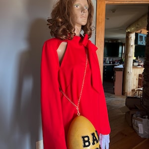May include: A bright red dress with a shoulder cut-out and long sleeves. A yellow egg-shaped purse with the text "BA EG" hangs from a silver chain. The mannequin wears white lace gloves.