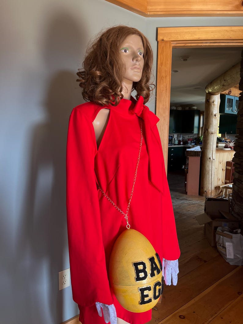 May include: A bright red dress with a shoulder cut-out and long sleeves. A yellow egg-shaped purse with the text "BA EG" hangs from a silver chain. The mannequin wears white lace gloves.