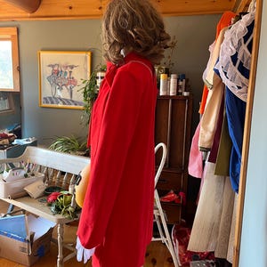May include: A vibrant red coat with white detailing and long sleeves. The coat is knee-length and has a classic design. The coat is displayed on a mannequin in a room with wooden floors and a bench.