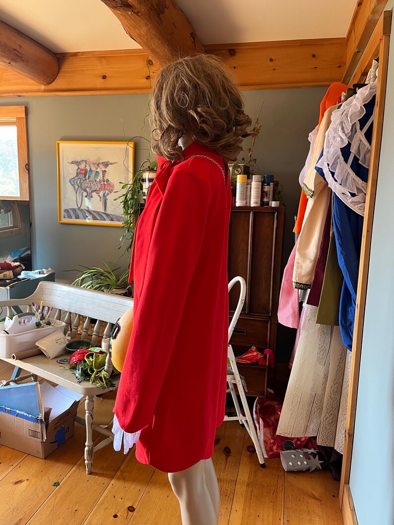 May include: A vibrant red coat with white detailing and long sleeves. The coat is knee-length and has a classic design. The coat is displayed on a mannequin in a room with wooden floors and a bench.