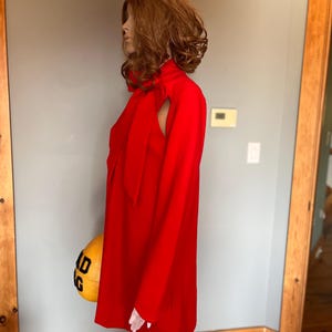 May include: A bright red dress with long sleeves and a high neckline. The dress has a distinctive shoulder cutout and a bow at the neck. The dress is displayed on a mannequin.