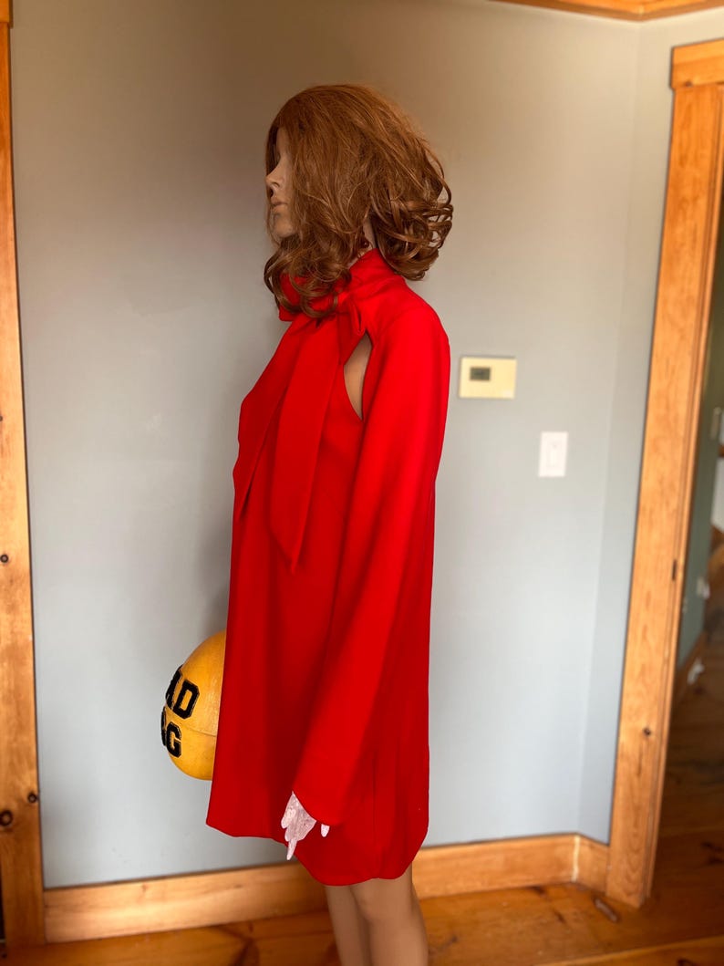 May include: A bright red dress with long sleeves and a high neckline. The dress has a distinctive shoulder cutout and a bow at the neck. The dress is displayed on a mannequin.