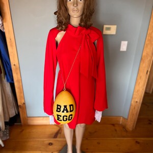 May include: A red dress with long sleeves and a bow detail, displayed on a mannequin. A yellow egg-shaped purse with the words "BAD EGG" in black hangs from the mannequin's shoulder. The mannequin stands on a dark green base.