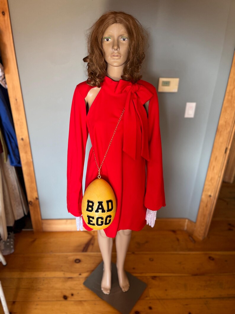 May include: A red dress with long sleeves and a bow detail, displayed on a mannequin. A yellow egg-shaped purse with the words "BAD EGG" in black hangs from the mannequin's shoulder. The mannequin stands on a dark green base.