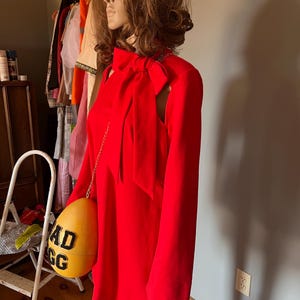 May include: A vibrant red dress featuring long sleeves and a bow at the neckline. The dress is accessorized with a yellow egg-shaped purse, displaying the text "AD EGG" in black. The mannequin is wearing white gloves.