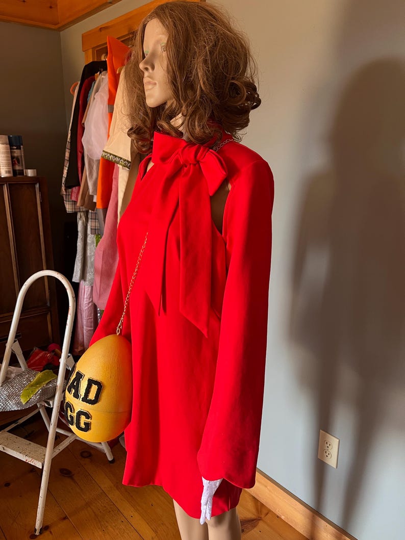 May include: A vibrant red dress featuring long sleeves and a bow at the neckline. The dress is accessorized with a yellow egg-shaped purse, displaying the text "AD EGG" in black. The mannequin is wearing white gloves.
