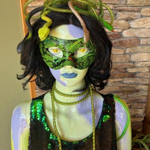 May include: A mannequin head and torso featuring a green snakeskin patterned eye mask, black hair, and a headpiece made of multiple green and brown plastic snakes. The mannequin wears a black sequined top and a long, thin, gold-toned beaded necklace.