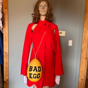 May include: A red dress with long sleeves and a high neckline. A yellow egg-shaped purse with the words "BAD EGG" in black is hanging from a gold chain. The mannequin is wearing white lace gloves.
