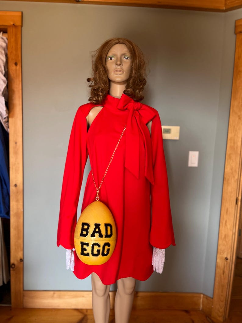 May include: A red dress with long sleeves and a high neckline. A yellow egg-shaped purse with the words "BAD EGG" in black is hanging from a gold chain. The mannequin is wearing white lace gloves.