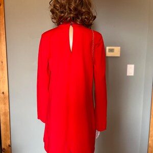 May include: A vibrant red dress featuring long sleeves and a keyhole back. The dress has a scalloped hem and a high neckline. A gold chain strap is visible. The dress is displayed on a mannequin.