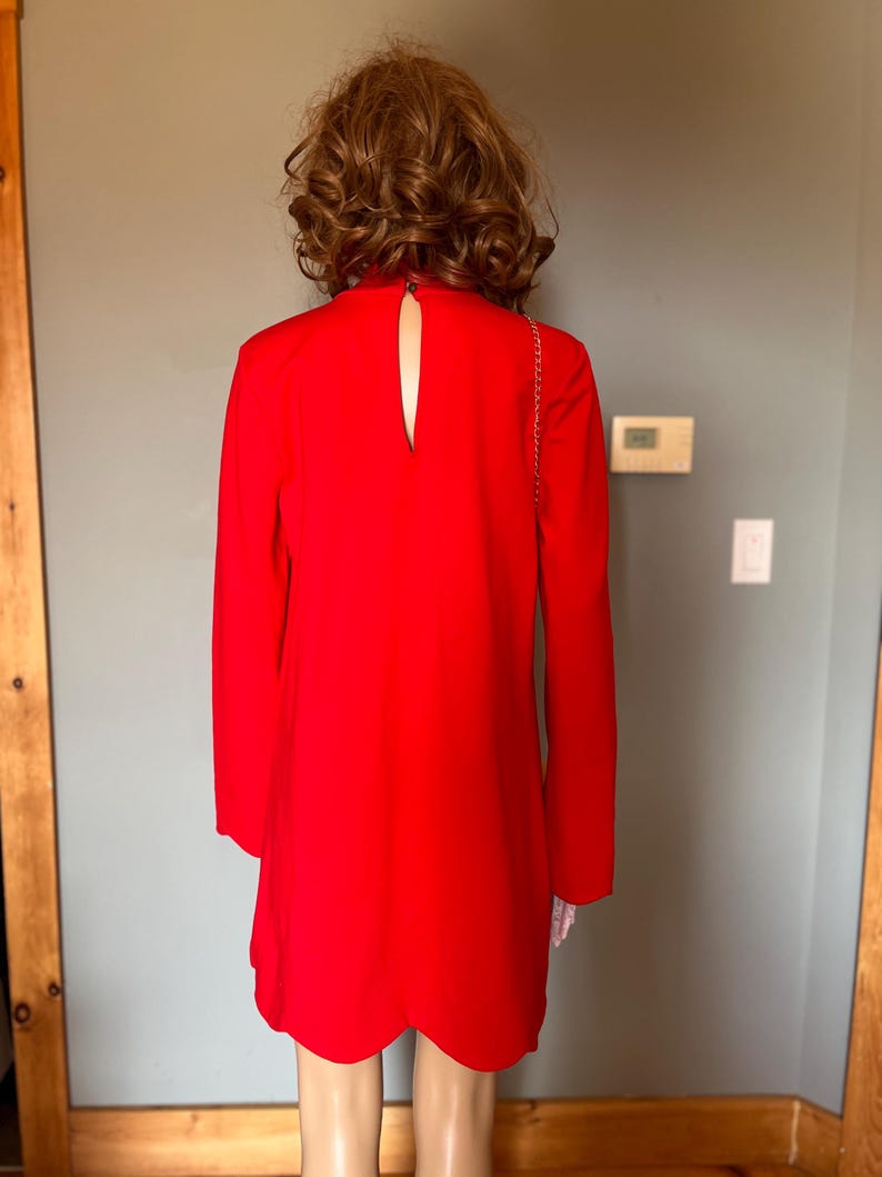 May include: A vibrant red dress featuring long sleeves and a keyhole back. The dress has a scalloped hem and a high neckline. A gold chain strap is visible. The dress is displayed on a mannequin.