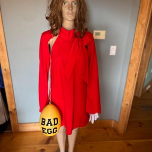 May include: A red dress with long sleeves and a bow detail, displayed on a mannequin. A yellow egg-shaped purse with the words "BAD EGG" in black hangs from the dress. The mannequin has a brown wig and is wearing white gloves.