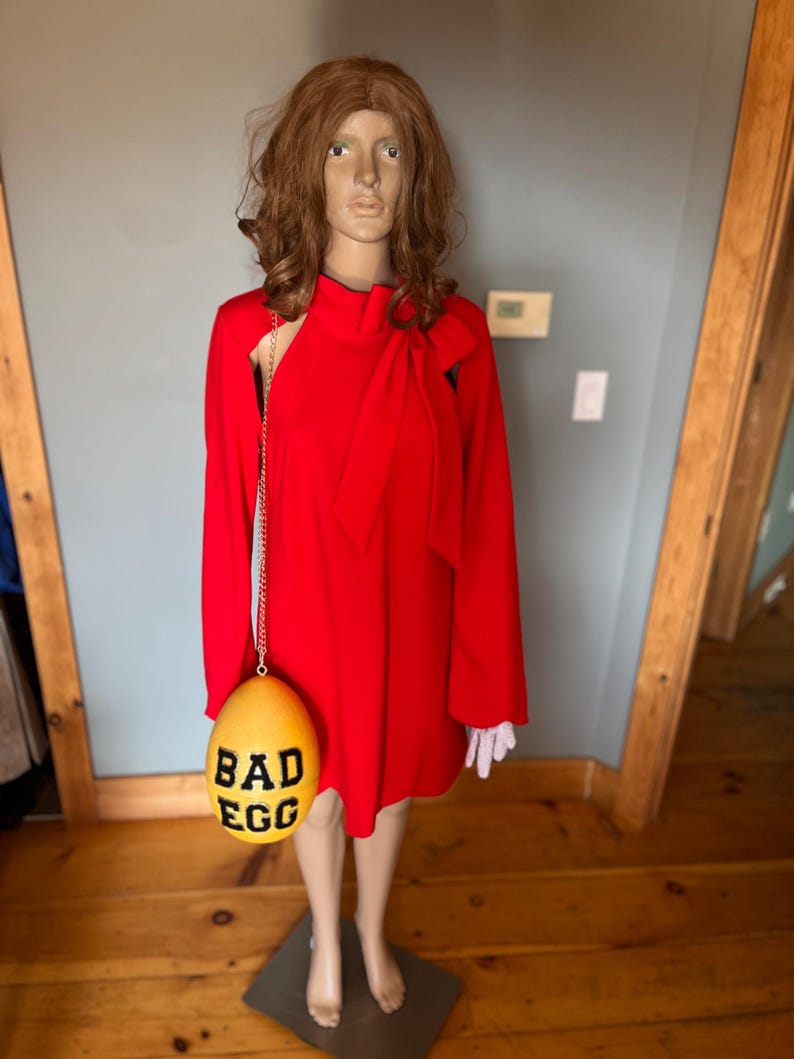 May include: A red dress with long sleeves and a bow detail, displayed on a mannequin. A yellow egg-shaped purse with the words "BAD EGG" in black hangs from the dress. The mannequin has a brown wig and is wearing white gloves.