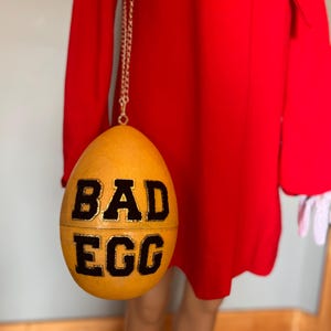 May include: A golden yellow egg-shaped handbag with the words "BAD EGG" in black lettering. The bag hangs from a silver chain. The bag is set against a bright red dress.