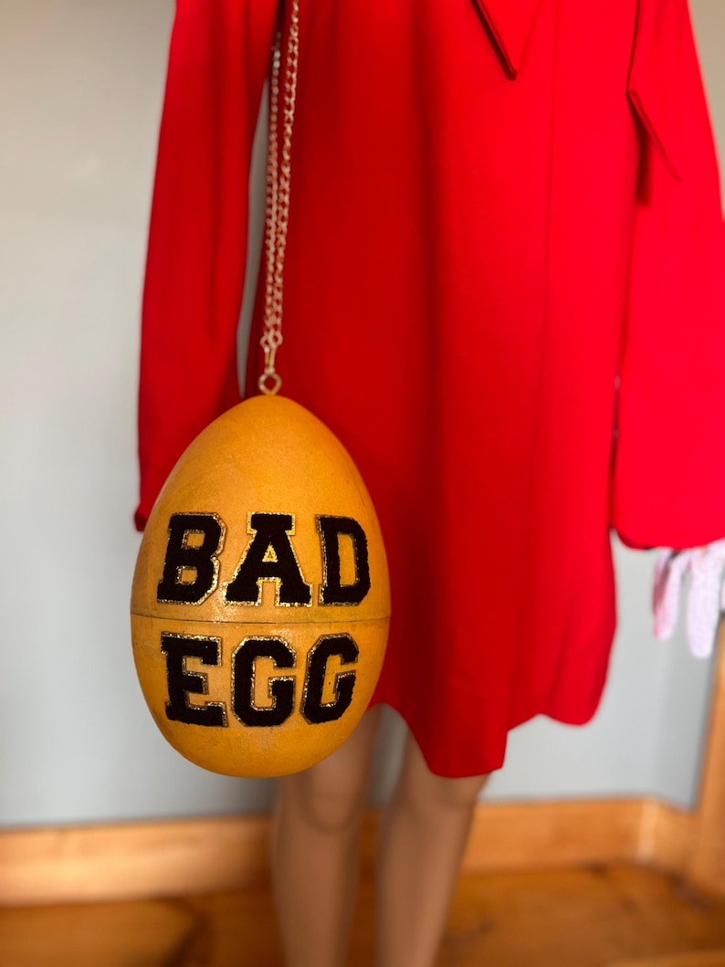 May include: A golden yellow egg-shaped handbag with the words "BAD EGG" in black lettering. The bag hangs from a silver chain. The bag is set against a bright red dress.
