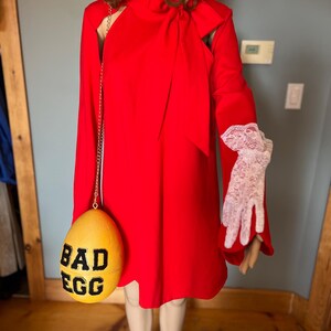 May include: A vibrant red dress with a high neckline and long sleeves. A yellow egg-shaped purse with the words "BAD EGG" in black is hanging from a gold chain. The mannequin is wearing white lace gloves.