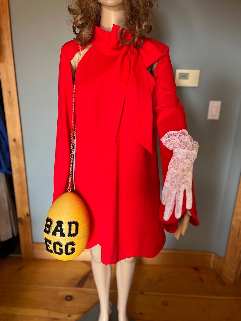 May include: A vibrant red dress with a high neckline and long sleeves. A yellow egg-shaped purse with the words "BAD EGG" in black is hanging from a gold chain. The mannequin is wearing white lace gloves.