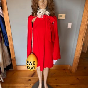 May include: A red dress with long sleeves and a bow detail, displayed on a mannequin. The dress is paired with a yellow egg-shaped purse that reads "BAD EGG" in black letters. The mannequin is wearing a white lace collar.