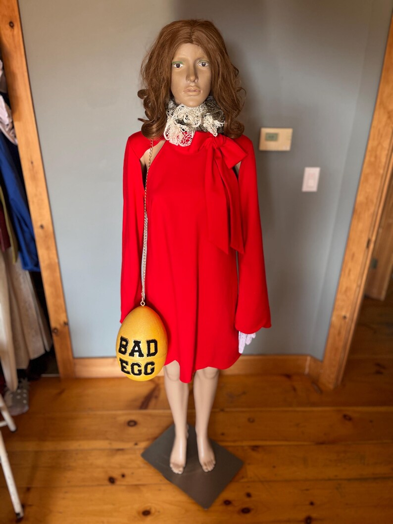May include: A red dress with long sleeves and a bow detail, displayed on a mannequin. The dress is paired with a yellow egg-shaped purse that reads "BAD EGG" in black letters. The mannequin is wearing a white lace collar.