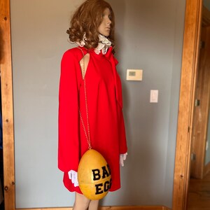 May include: A mannequin in a vibrant red dress, accessorised with a gold chain and a yellow egg-shaped purse. The purse features the words "BAD EGG" in black. The dress has a high collar with white lace trim and a cut-out shoulder.