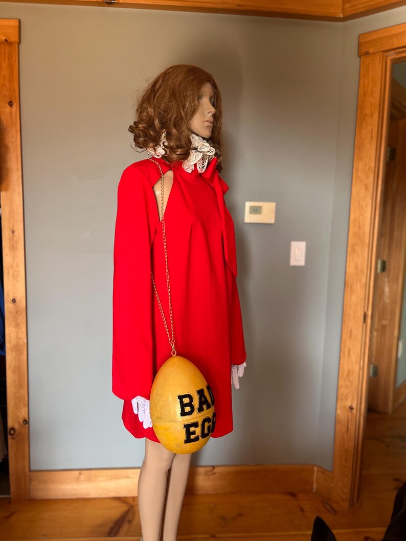 May include: A mannequin in a vibrant red dress, accessorised with a gold chain and a yellow egg-shaped purse. The purse features the words "BAD EGG" in black. The dress has a high collar with white lace trim and a cut-out shoulder.