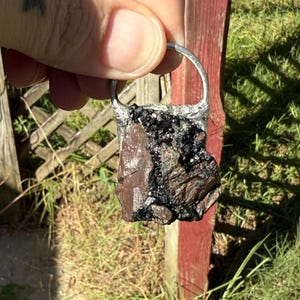 May include: A pendant featuring a rough-cut, dark brown and black stone set in a silver-colored metal frame. The stone has a textured surface with visible crystalline structures. The pendant is held up by a silver loop.