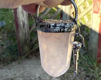 May include: A rose quartz crystal pendant with a silver-colored metal cap and bail. The pendant has a small, silver-colored charm attached. The crystal is a pale pink color and is held up by a hand.