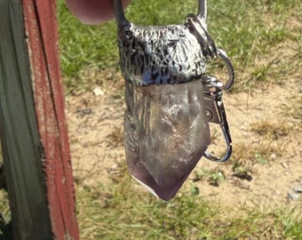 May include: A raw amethyst crystal pendant with a silver-colored metal bail and clasp. The crystal is a translucent purple color and is held up against a background of green grass and a red wooden post.