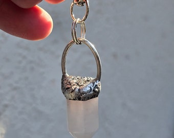 May include: A silver keychain with a clear quartz crystal pendant. The crystal is shaped like a point and is attached to a silver ring. The keychain has a silver clasp.