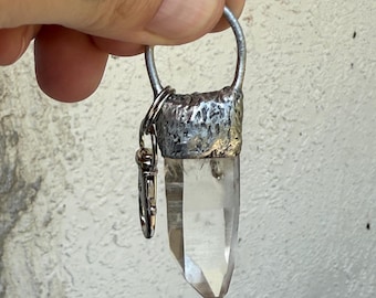 May include: Clear quartz crystal pendant with a silver-colored metal cap and ring. A small silver-colored clasp is attached to the ring. The crystal is a transparent, elongated shape with a pointed tip.