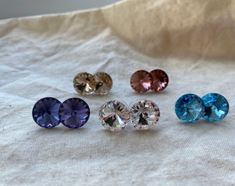 May include: A set of six pairs of round crystal earrings in various colors, including clear, light blue, purple, and pink. The earrings are displayed on a white fabric surface.
